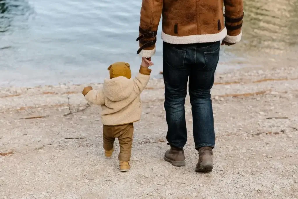 Parent walking hand-in-hand with toddler on the beach.